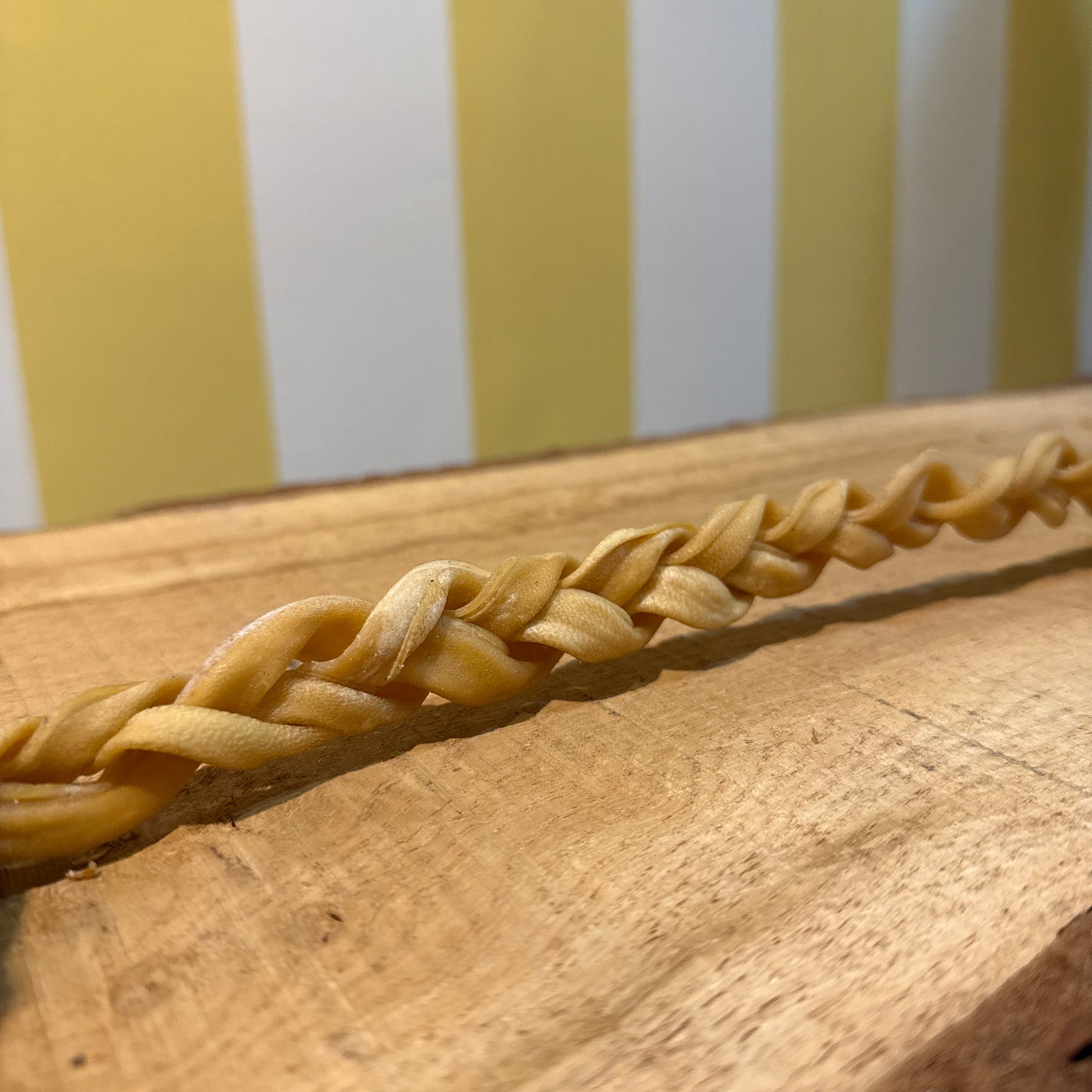 A close-up of a braided strand resembling The Tetbury Pet Shop & Grooming's Braided Lamb Chew 30cm for dogs, shown on wood with a yellow and white striped background.