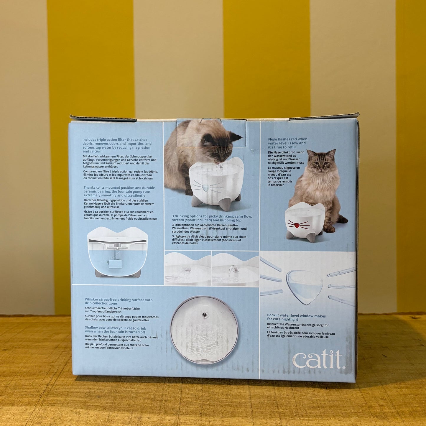 A box for The Tetbury Pet Shop & Grooming’s Catit Pixi drinking fountain, showing a cat and the fountain on a yellow-striped background. Features include an ergonomic design and triple action filter to support your pet’s health.