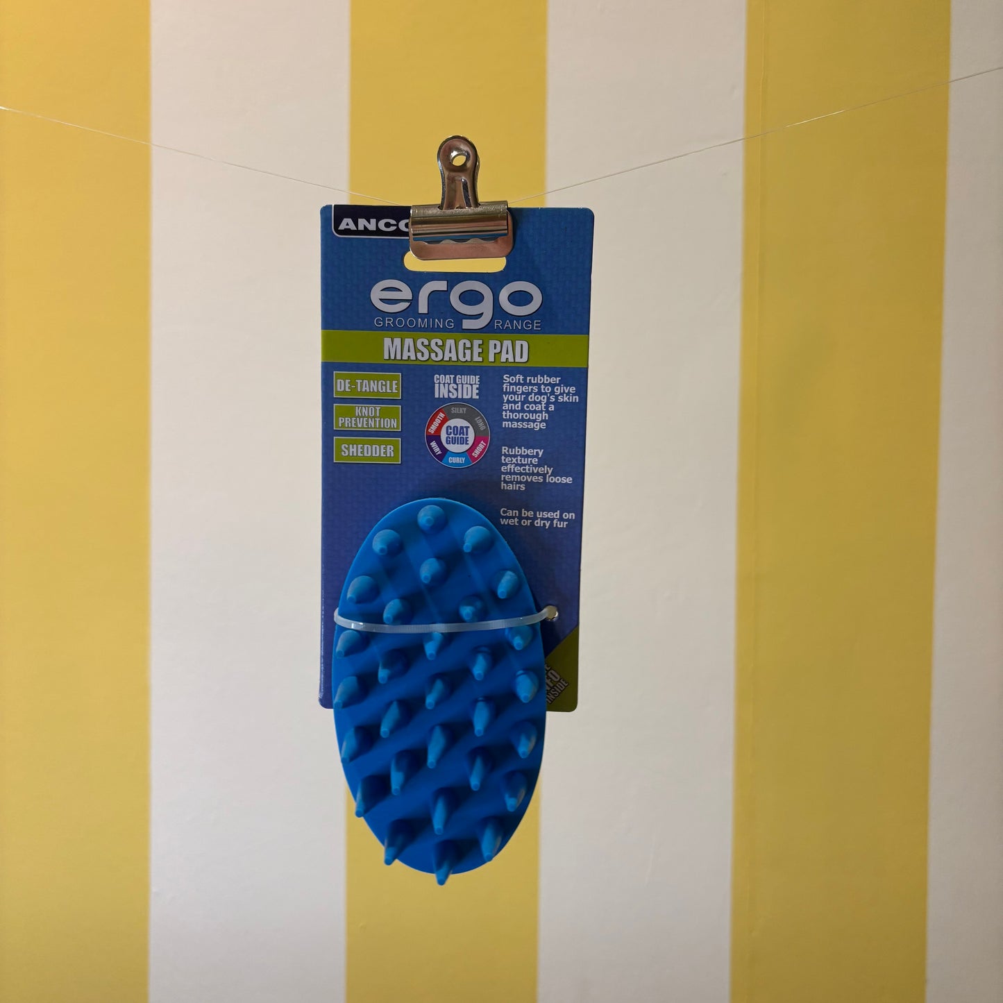 The ERGO Massage Pad by The Tetbury Pet Shop & Grooming, in blue and packaged, hangs on a clip before a yellow and white striped backdrop—ideal for gentle dog grooming.
