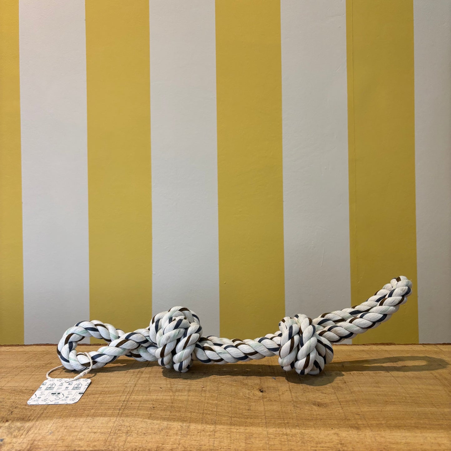 The Tetbury Pet Shop & Grooming’s Duo Knot Rope chew toy with tag sits on a wooden surface by a yellow and white striped wall—perfect toy for your dog’s dental health.