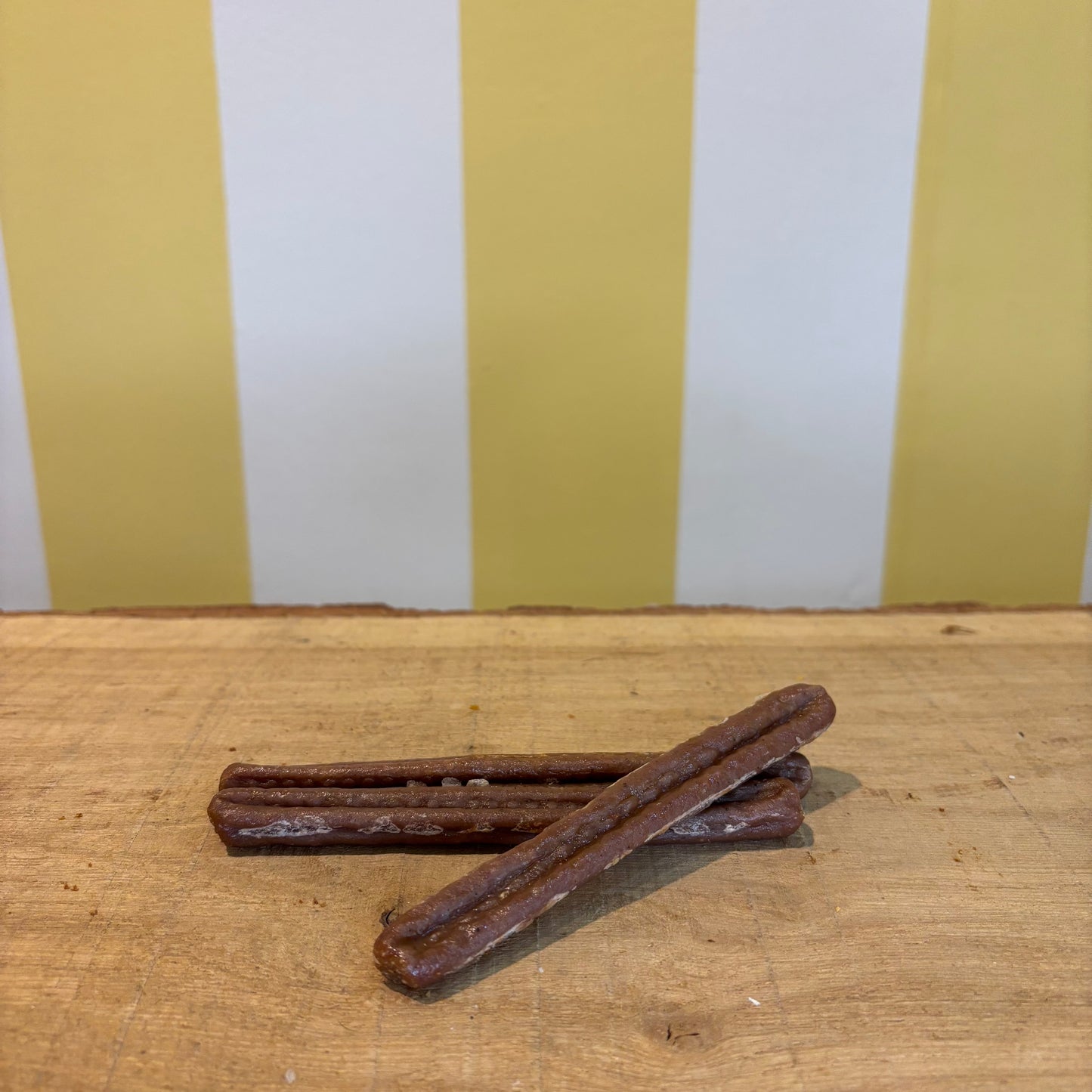 Two brown Sausages with textured surfaces are crossed on a wooden surface, in front of a yellow and white striped wall.