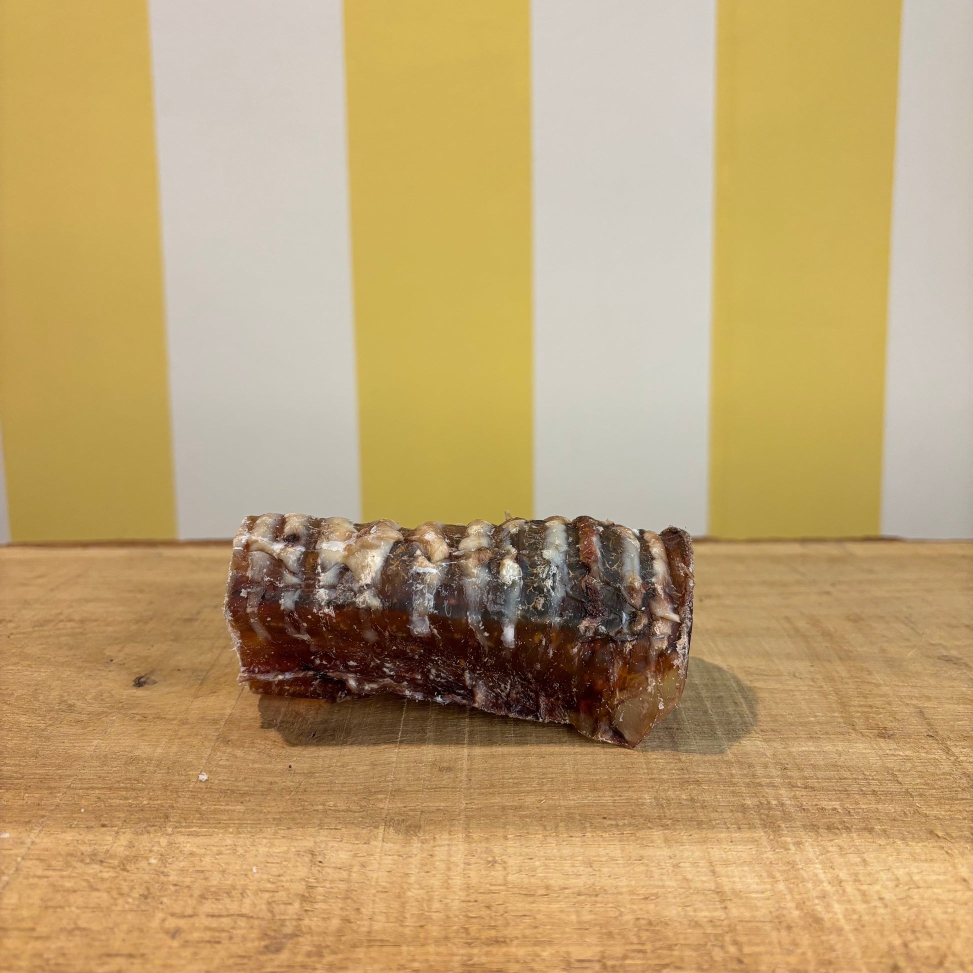 A single Beef Trachea chew, known for supporting joint health, sits on a wooden surface with visible marbling and texture, set against a yellow and white striped background.