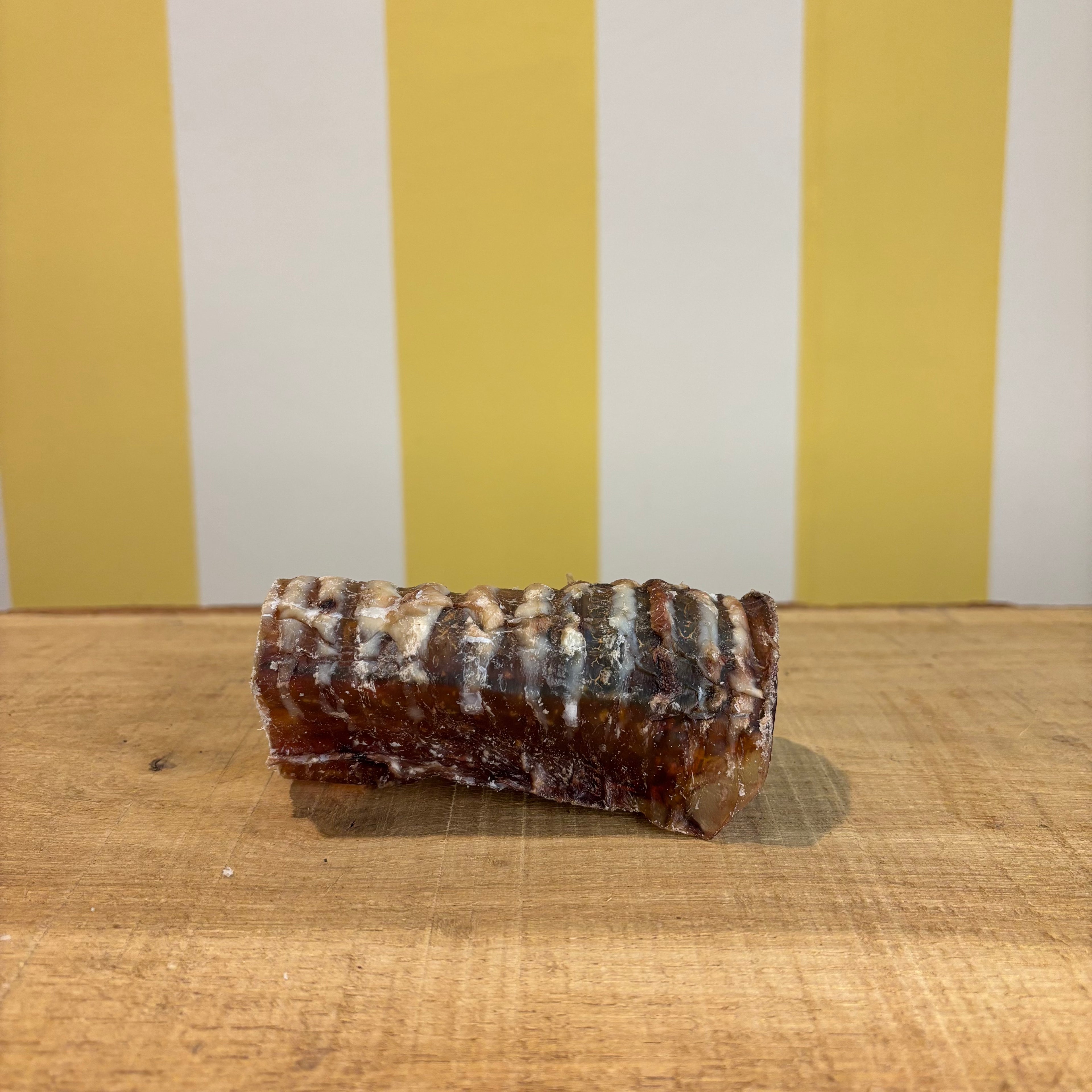 A single Beef Trachea chew, known for supporting joint health, sits on a wooden surface with visible marbling and texture, set against a yellow and white striped background.