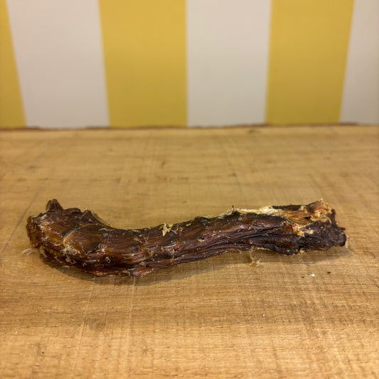 A single Duck Neck dog treat, which is grain free, rests on a wooden surface with a yellow and white striped background.