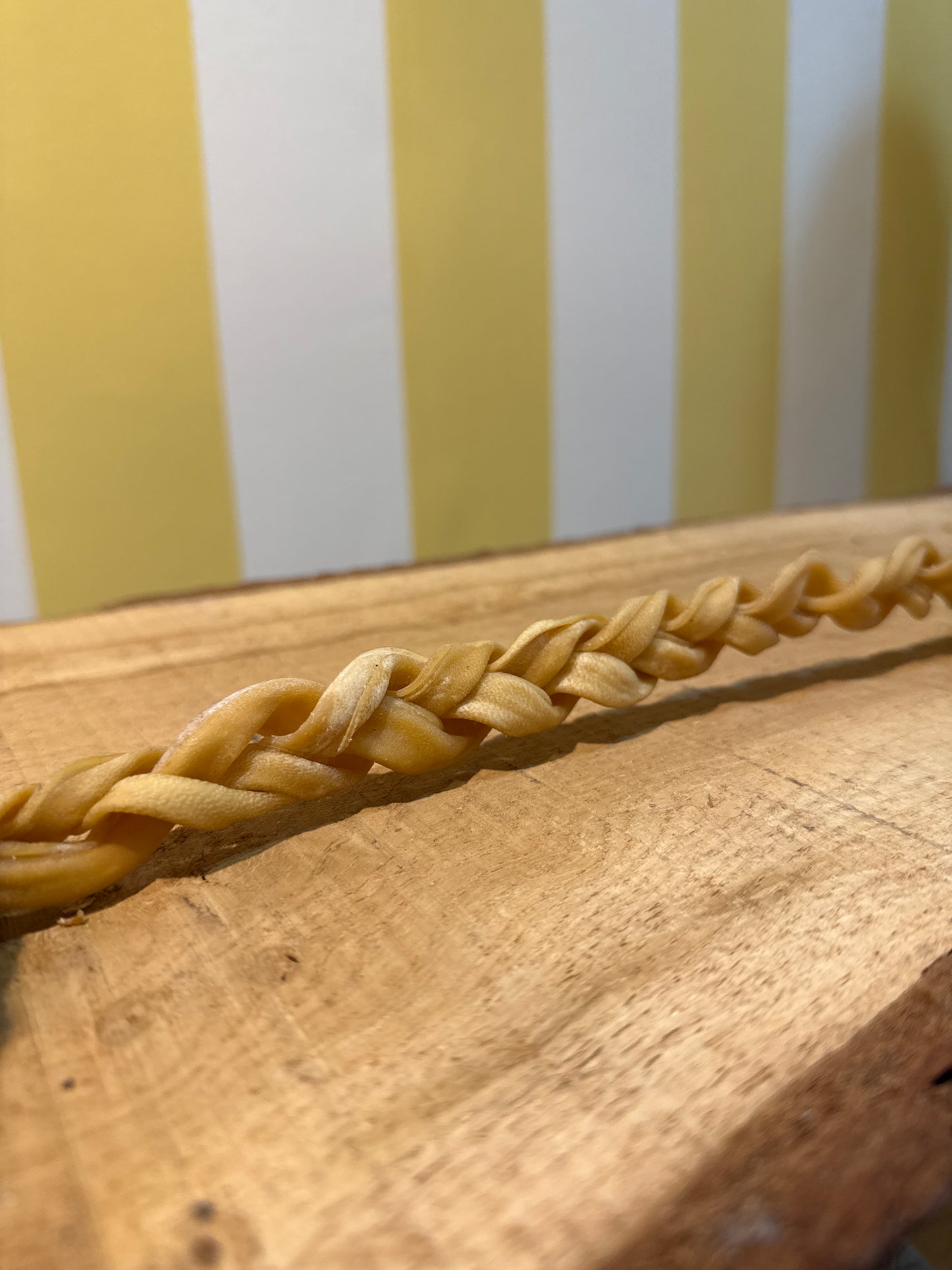 A close-up of a braided strand resembling The Tetbury Pet Shop & Grooming's Braided Lamb Chew 30cm for dogs, shown on wood with a yellow and white striped background.