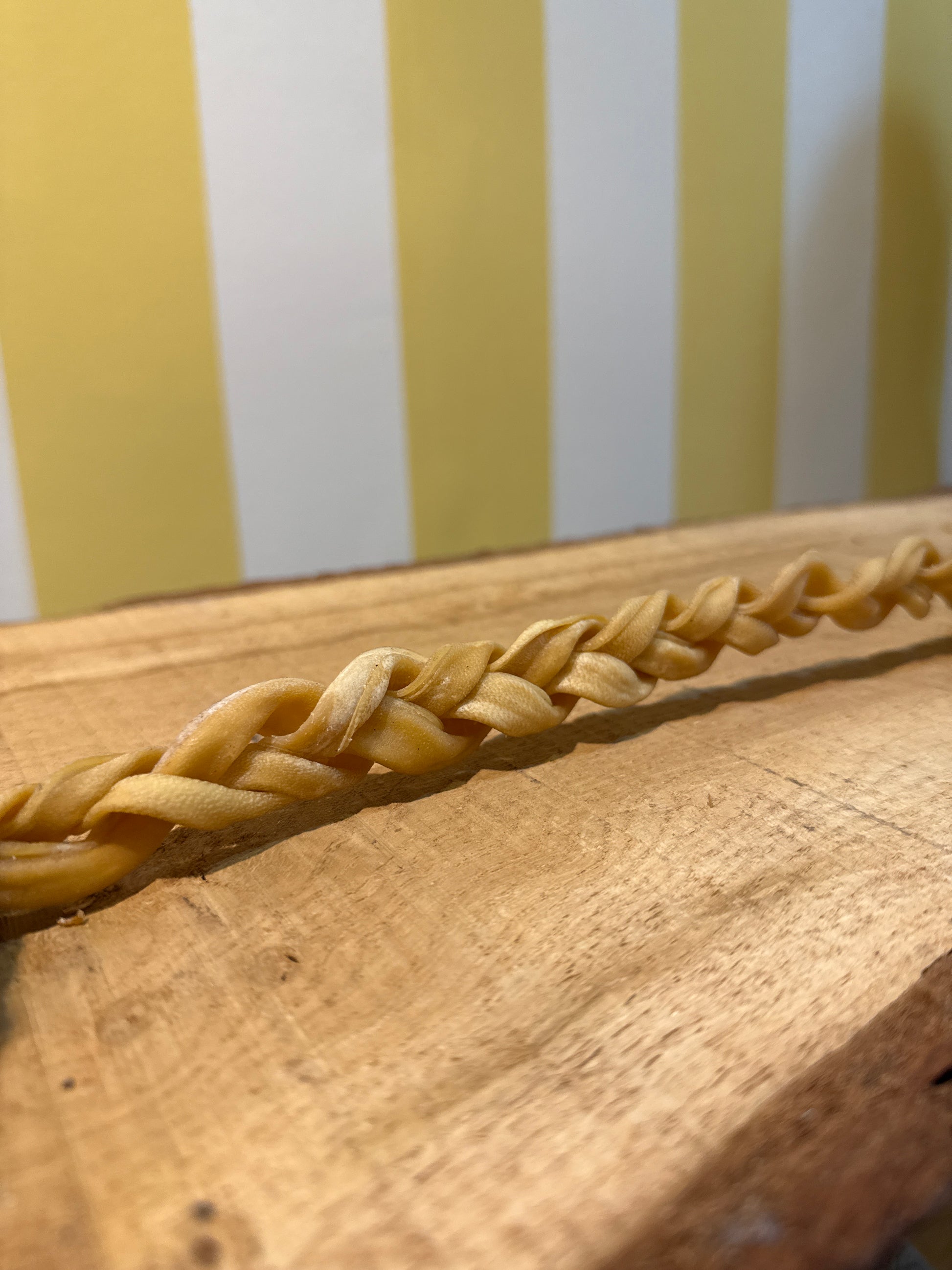 A close-up of a braided strand resembling The Tetbury Pet Shop & Grooming's Braided Lamb Chew 30cm for dogs, shown on wood with a yellow and white striped background.