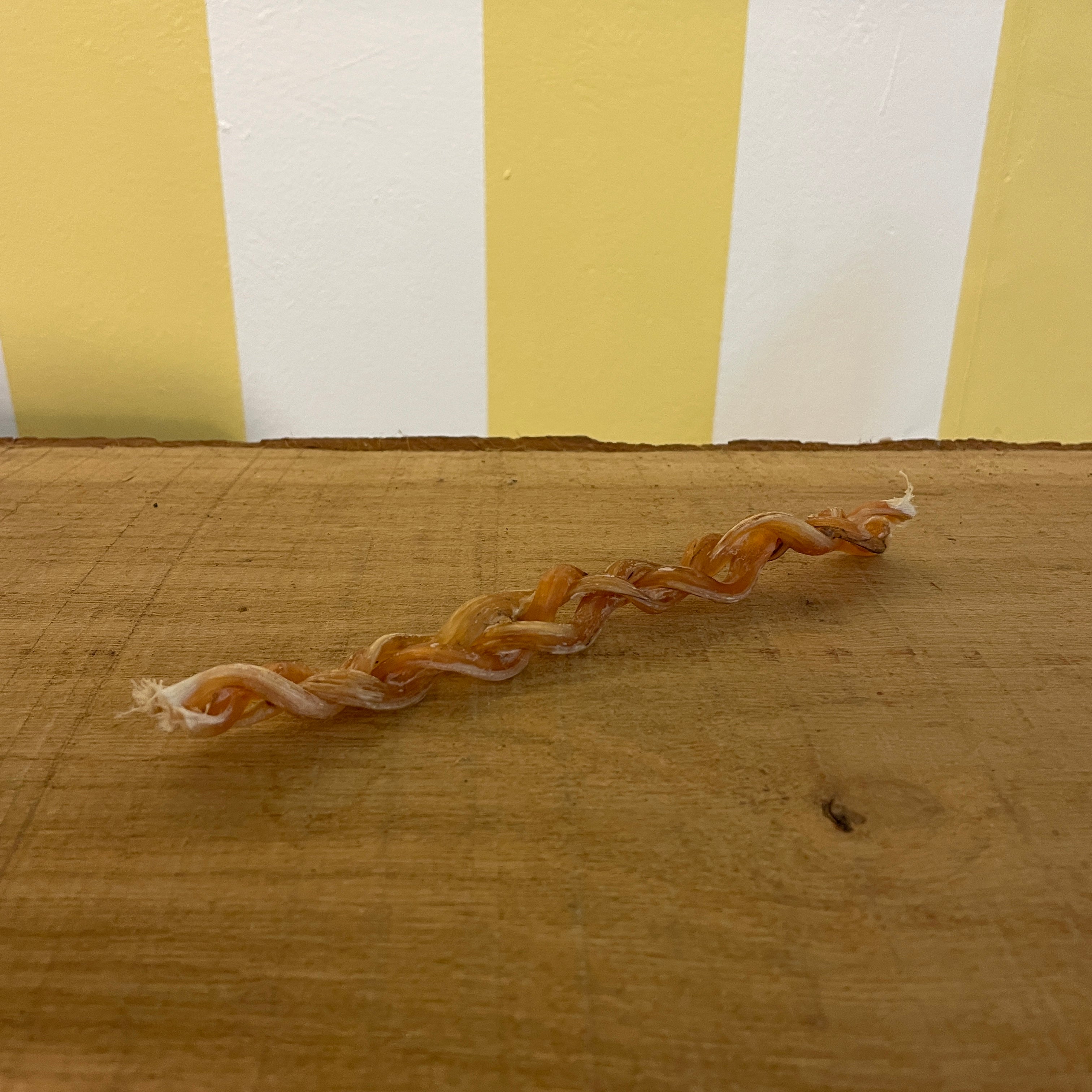A close-up image of a braided ostrich tendon chew on a wooden surface with a striped yellow and white background.