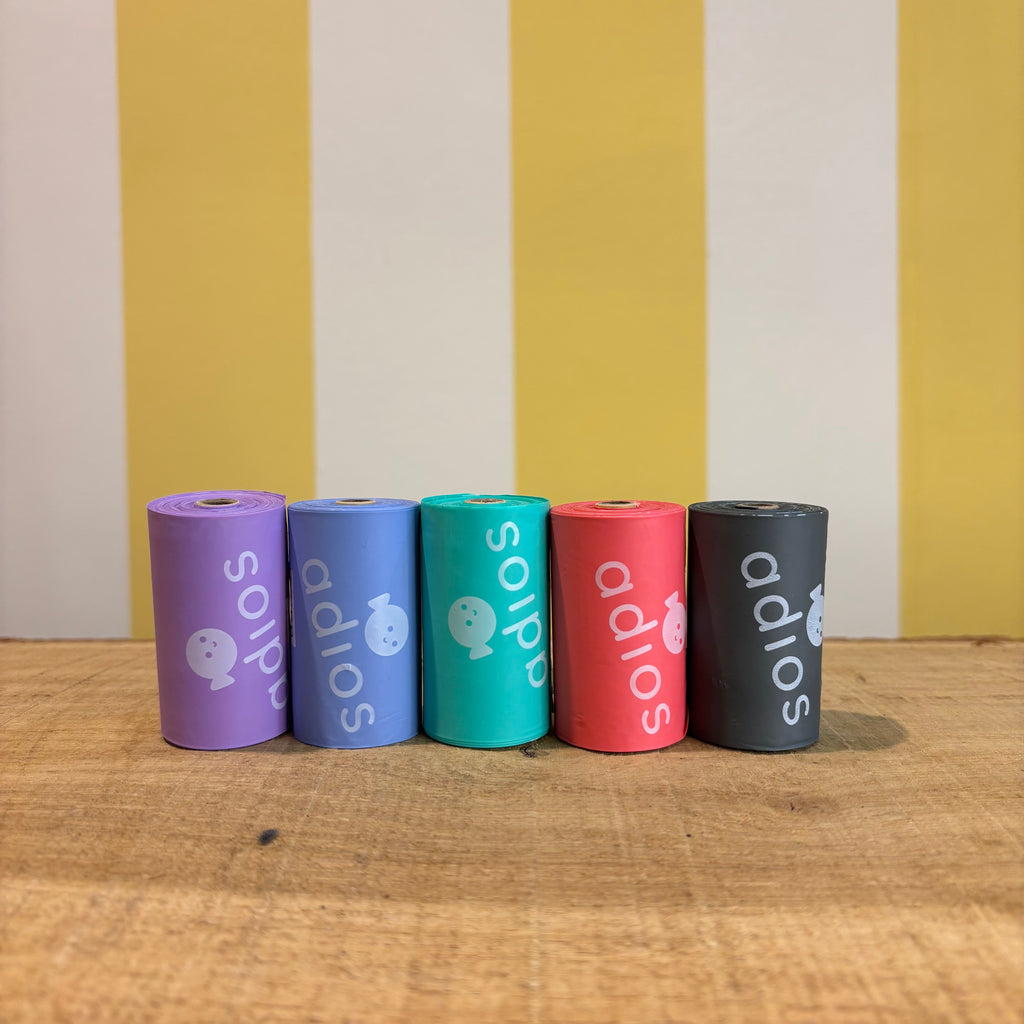 Five colorful rolls of Adios Poo Bags compostable dog waste bags—purple, blue, turquoise, coral, and black—stand upright on a wooden surface with a yellow and white striped wall in the background.