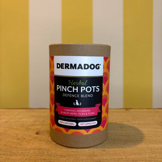 Grooming’s DermaDog Herbal Pinch Pots sits on a wooden surface, with a yellow and white striped background.