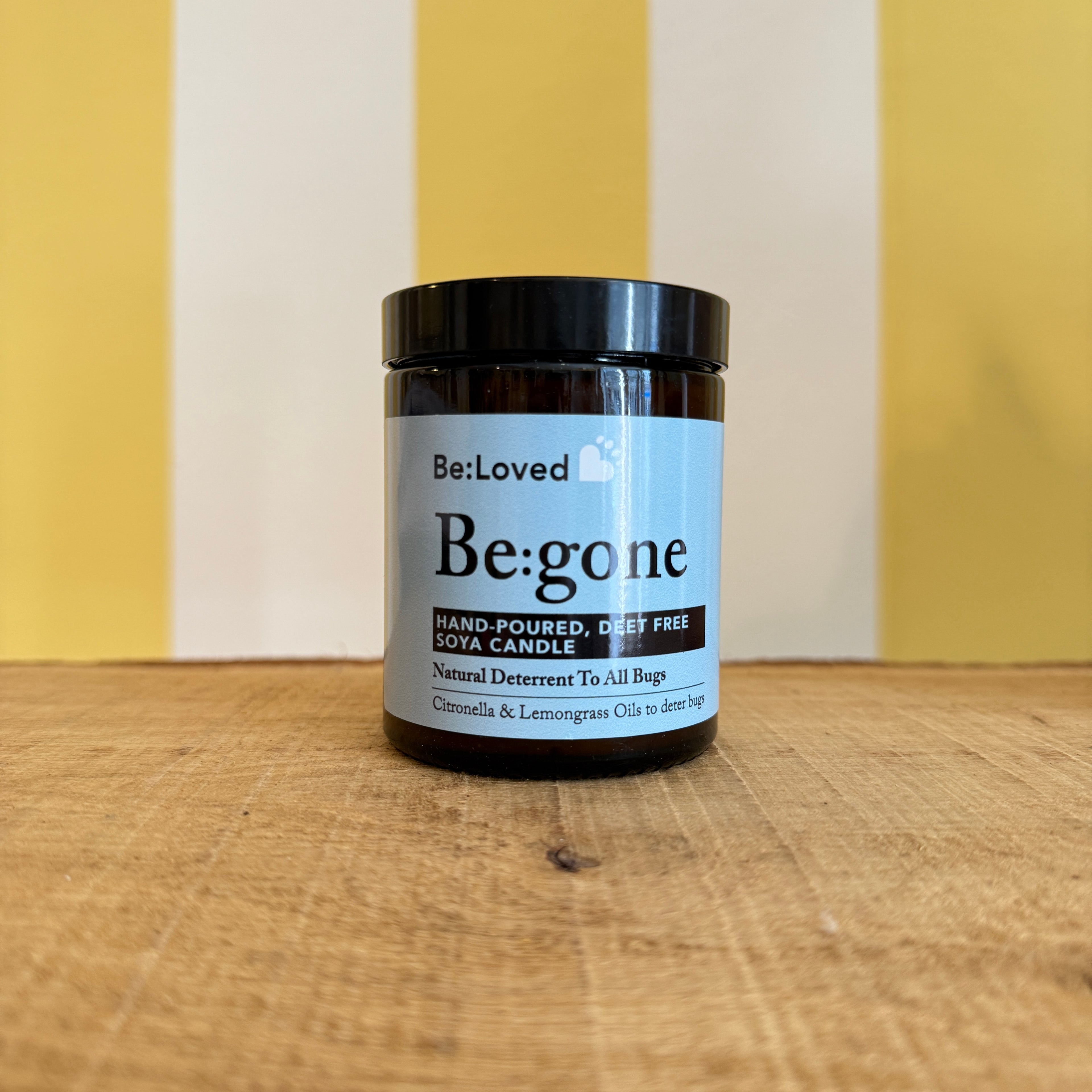 A jar labeled Be:Loved Candle by The Tetbury Pet Shop & Grooming, an odour eliminating soy candle, sits on a wooden surface against a yellow and white striped background.