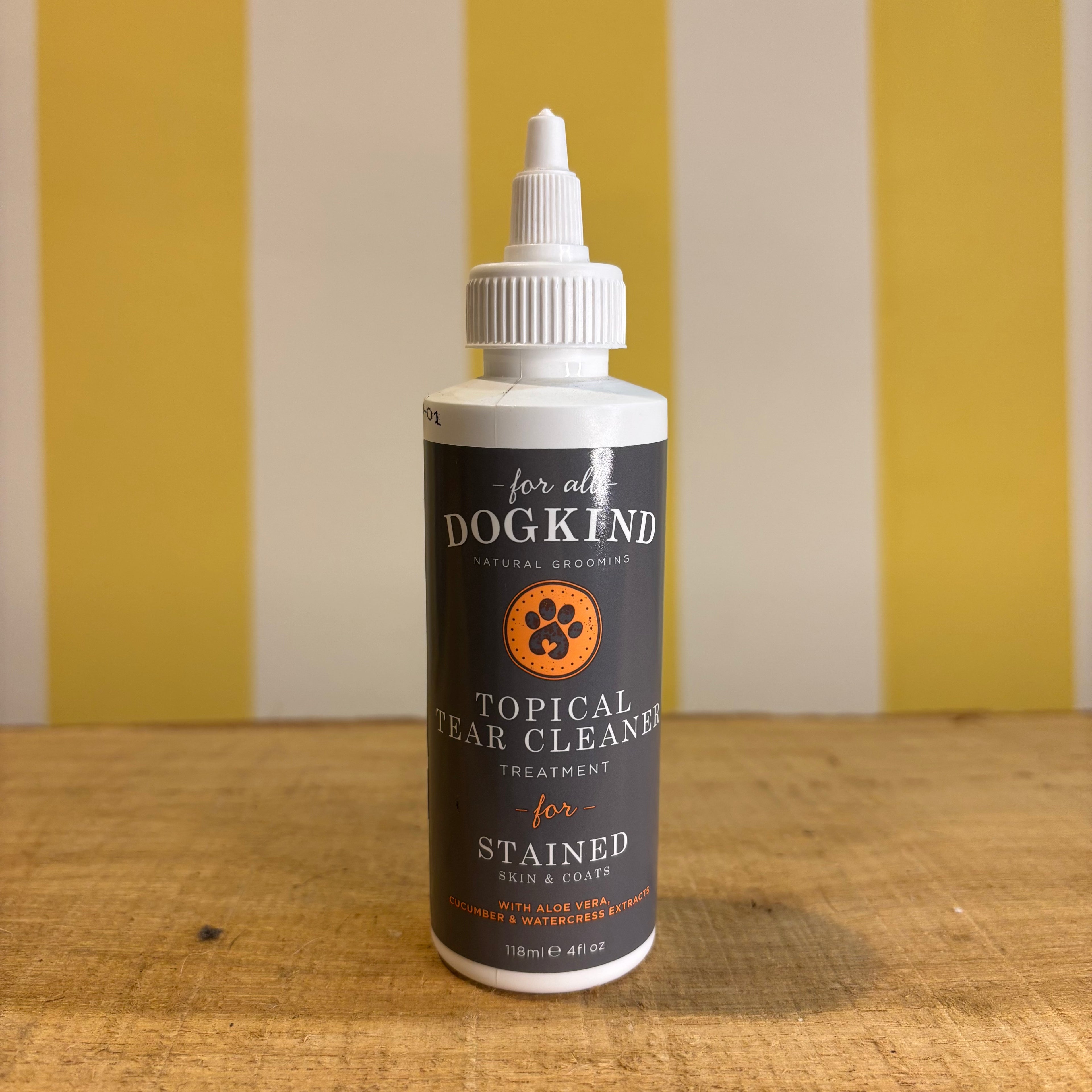 Three bottles of For all Dogkind dog shampoo by The Tetbury Pet Shop & Grooming, made with essential oils, are arranged on a wooden surface in front of a yellow and white striped background.