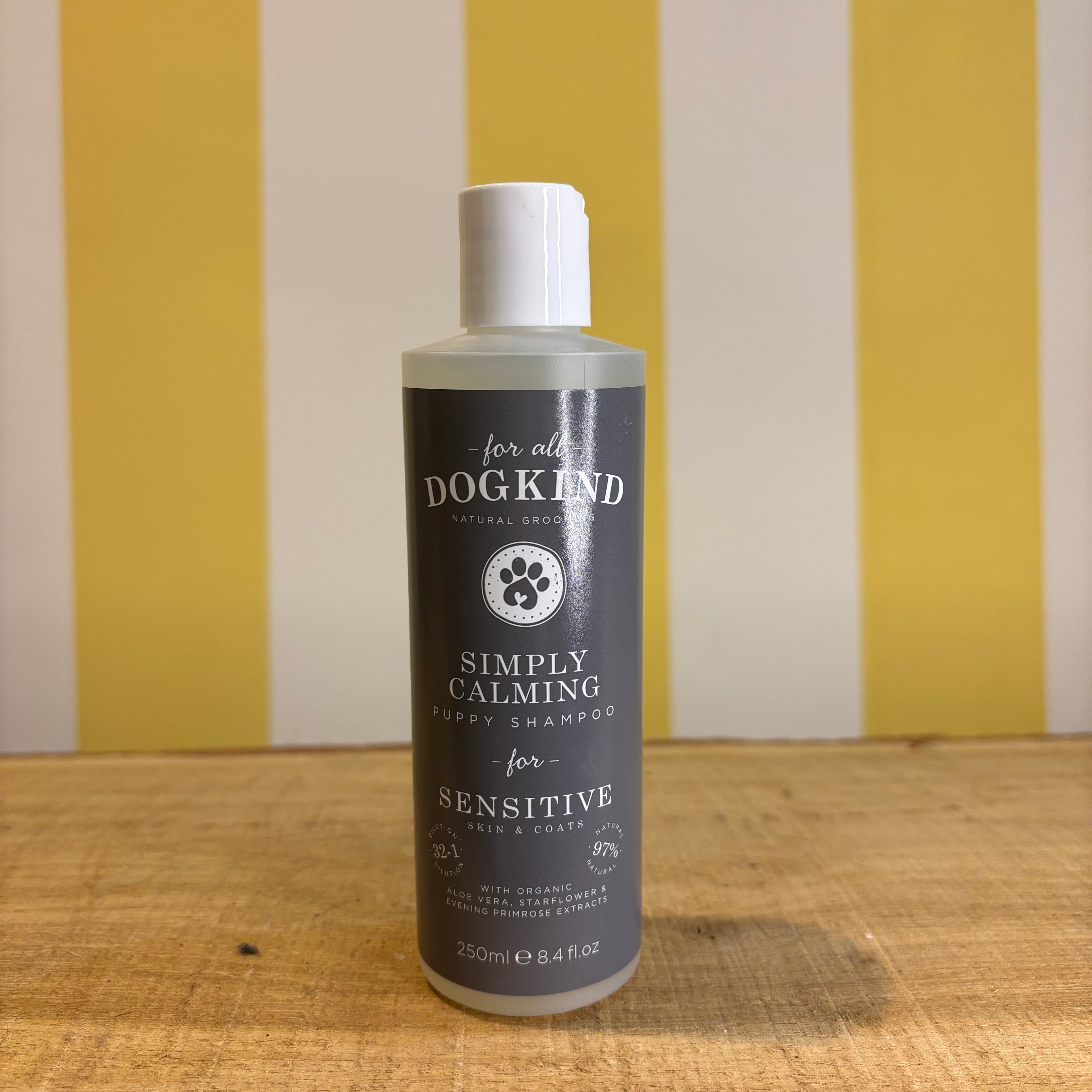 Three bottles of For all Dogkind dog shampoo by The Tetbury Pet Shop & Grooming, made with essential oils, are arranged on a wooden surface in front of a yellow and white striped background.