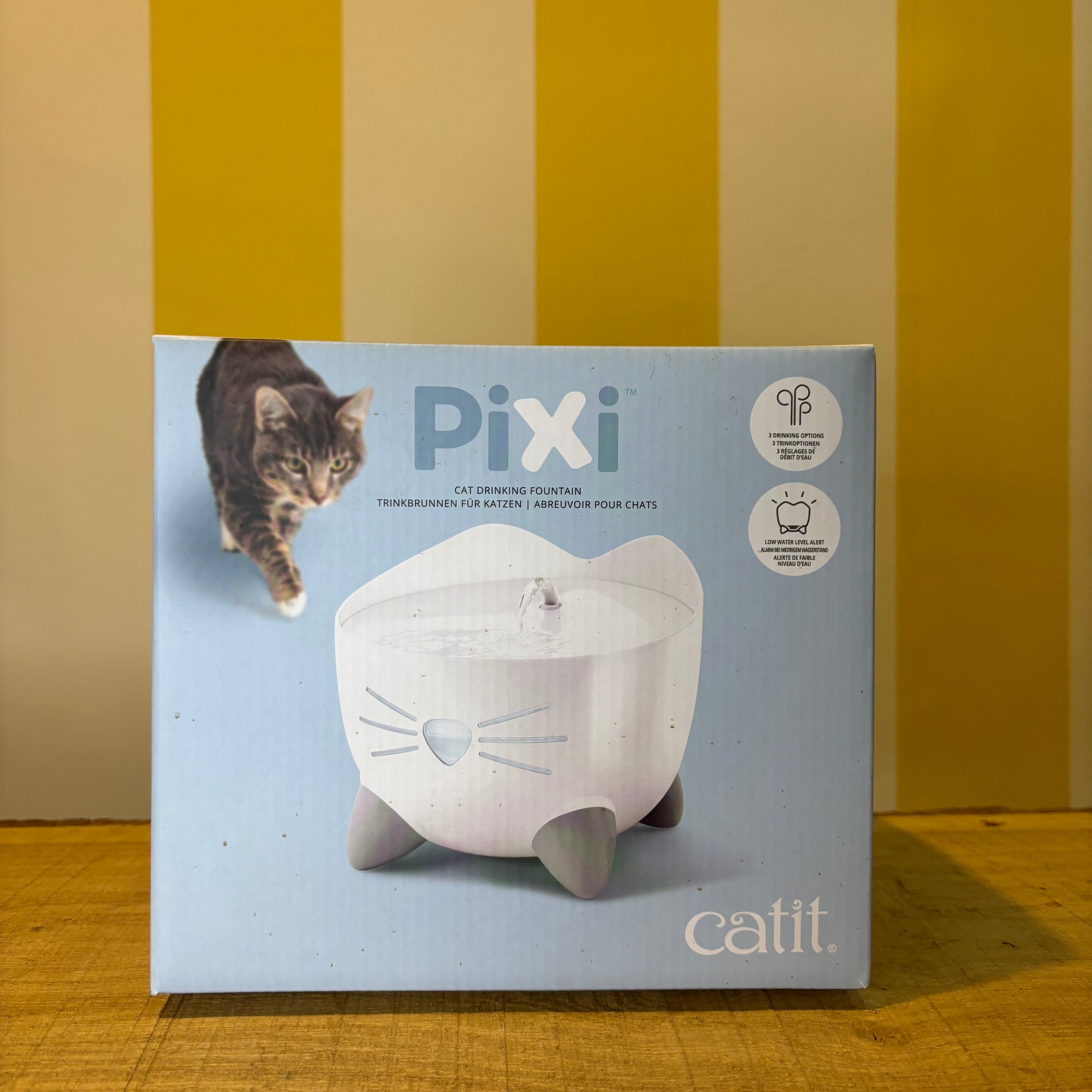 A box for The Tetbury Pet Shop & Grooming’s Catit Pixi drinking fountain, showing a cat and the fountain on a yellow-striped background. Features include an ergonomic design and triple action filter to support your pet’s health.