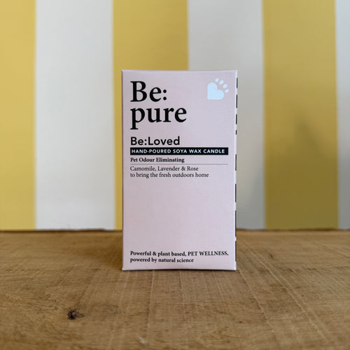 A jar labeled Be:Loved Candle by The Tetbury Pet Shop & Grooming, an odour eliminating soy candle, sits on a wooden surface against a yellow and white striped background.