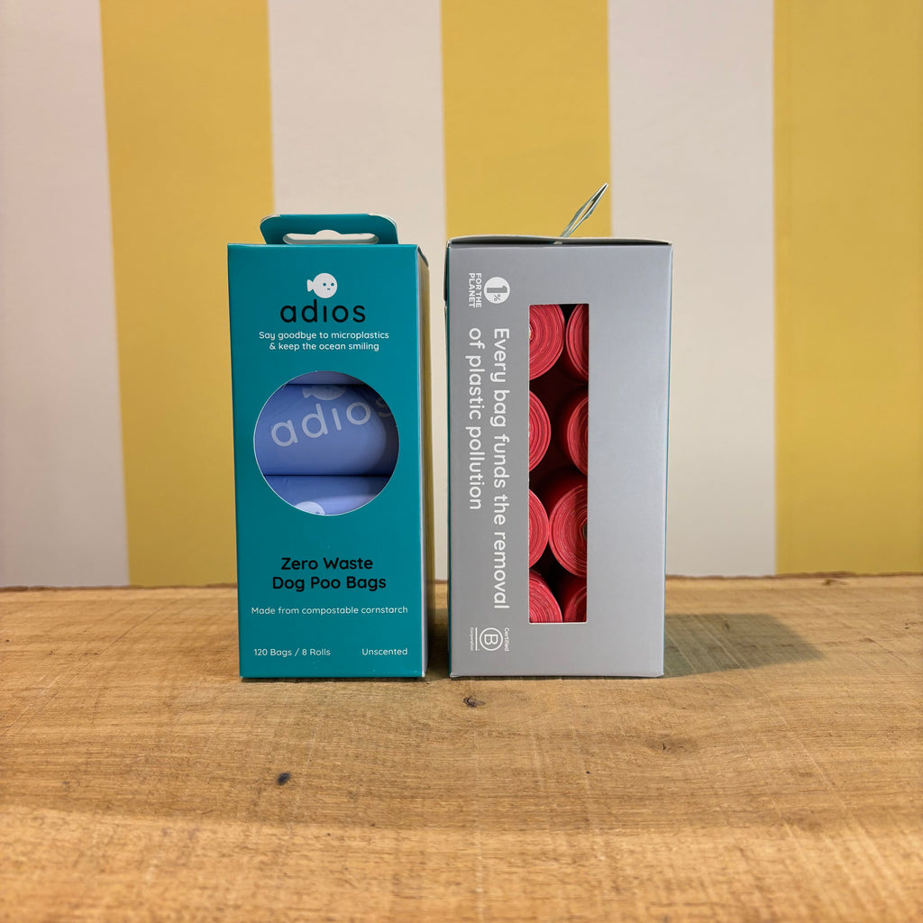 Two boxes of Adios Poo Bags on a wooden surface with a yellow and white striped background—one blue box labeled Zero Waste Dog Poo Bags, and one gray box displaying pink, biodegradable dog bags inside.