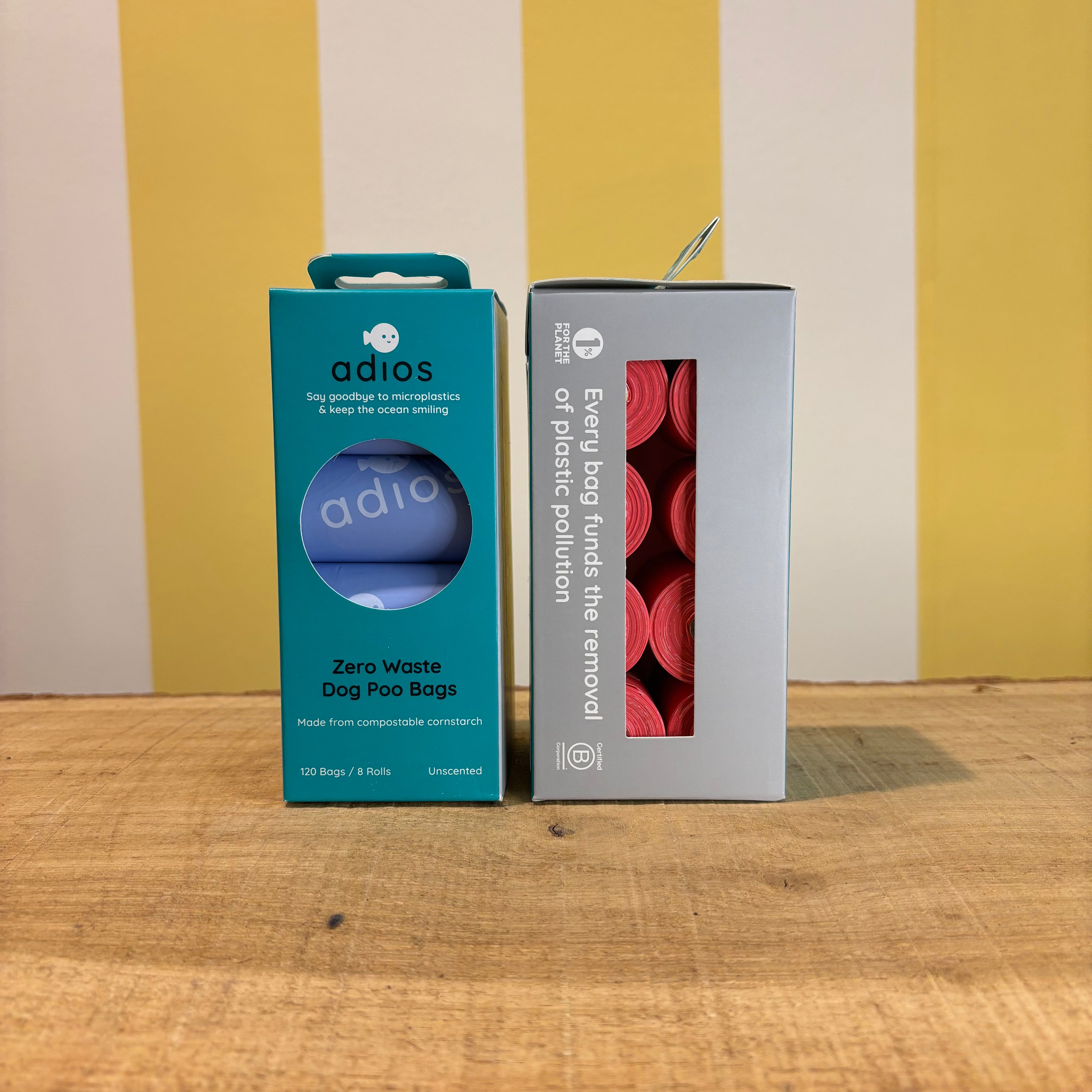 Two boxes of Adios Poo Bags on a wooden surface with a yellow and white striped background—one blue box labeled Zero Waste Dog Poo Bags, and one gray box displaying pink, biodegradable dog bags inside.