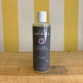 Three bottles of For all Dogkind dog shampoo by The Tetbury Pet Shop & Grooming, made with essential oils, are arranged on a wooden surface in front of a yellow and white striped background.