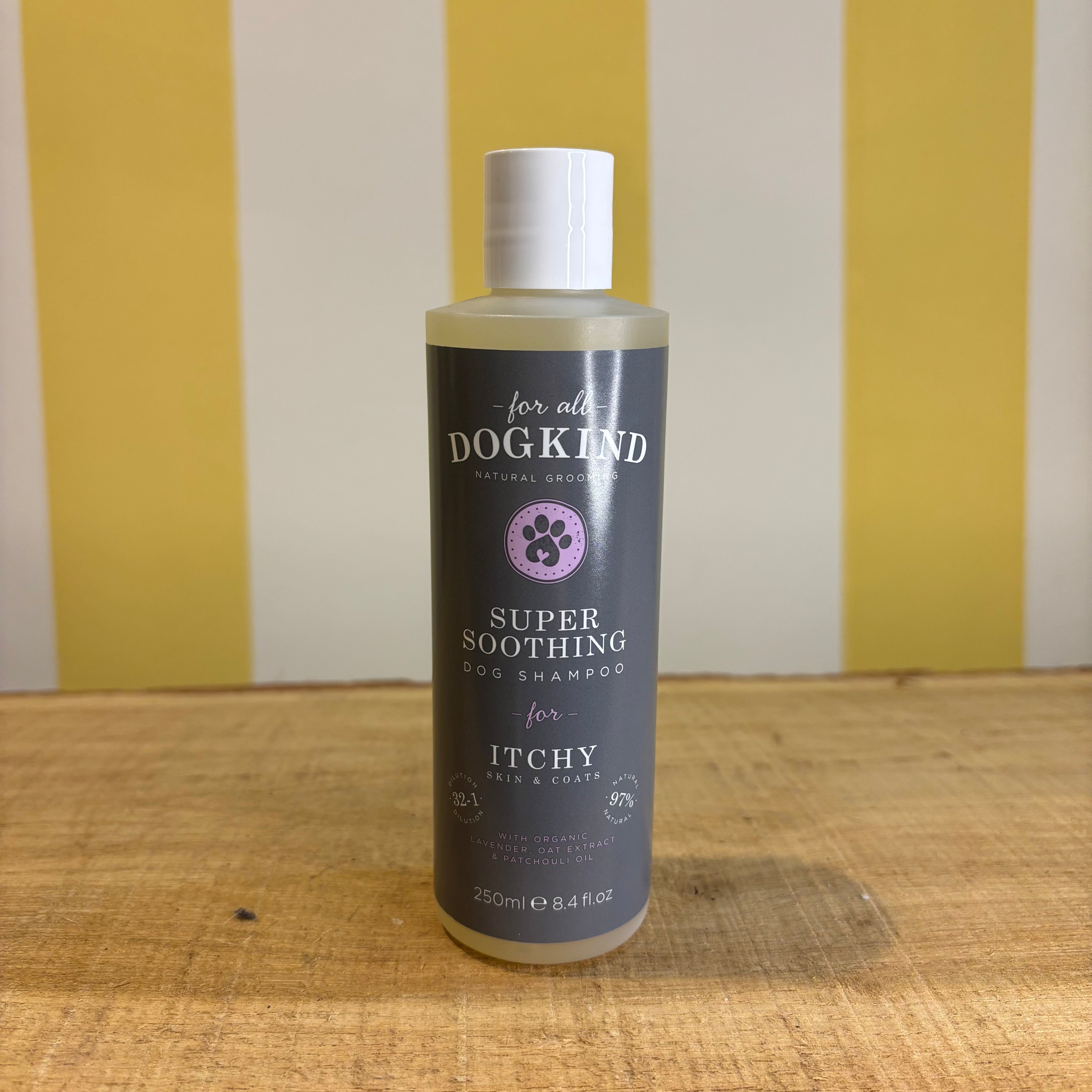 Three bottles of For all Dogkind dog shampoo by The Tetbury Pet Shop & Grooming, made with essential oils, are arranged on a wooden surface in front of a yellow and white striped background.