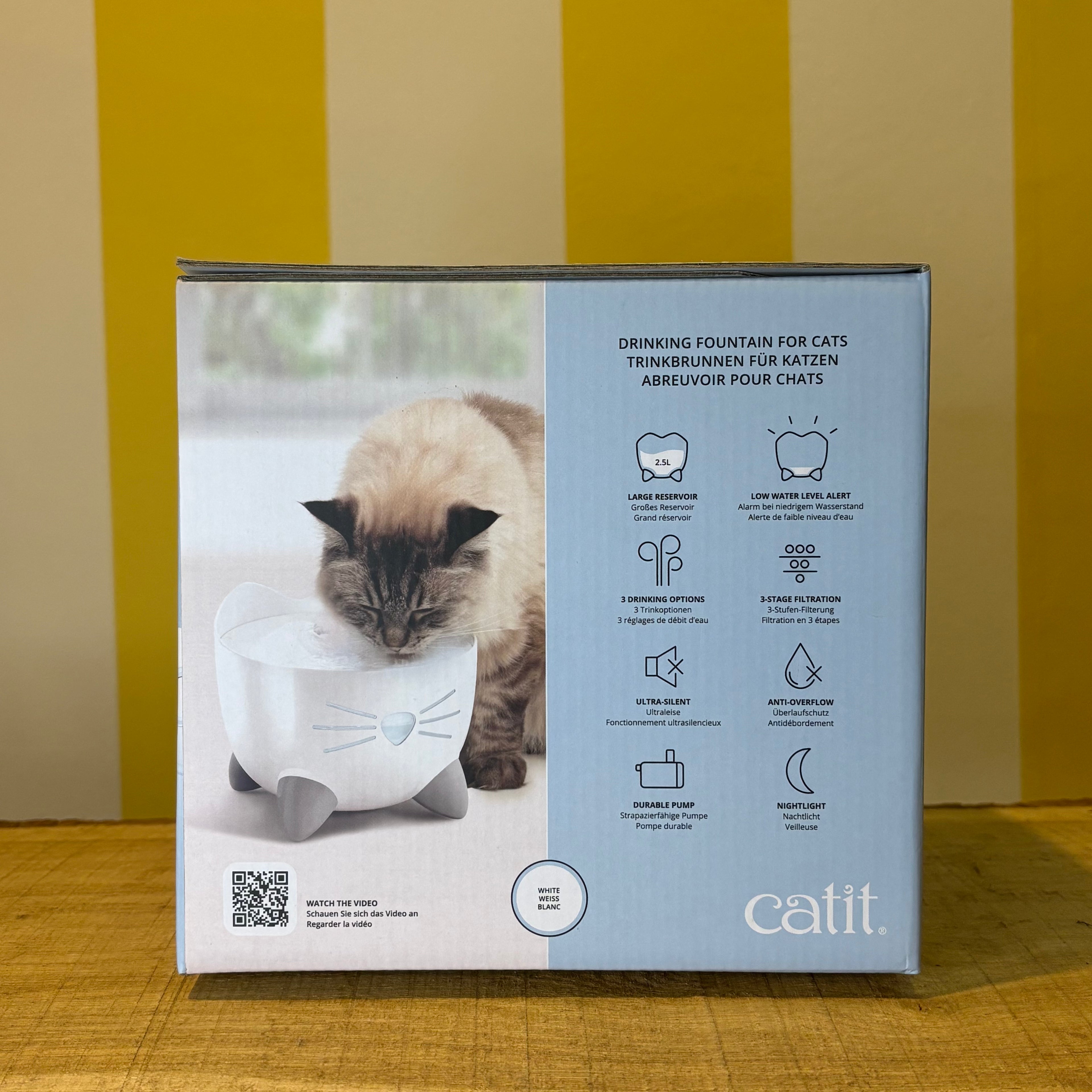 A box for The Tetbury Pet Shop & Grooming’s Catit Pixi drinking fountain, showing a cat and the fountain on a yellow-striped background. Features include an ergonomic design and triple action filter to support your pet’s health.