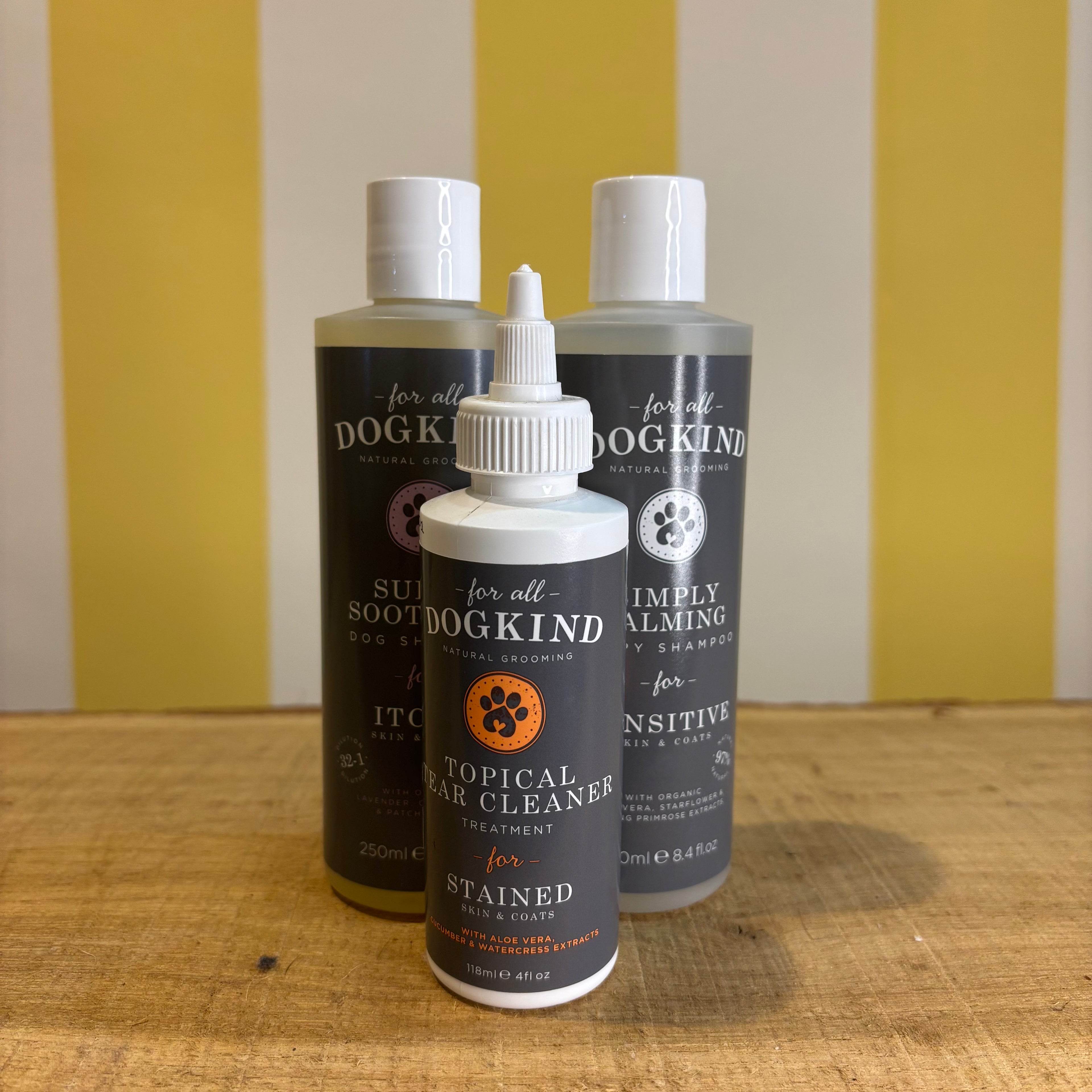 Three bottles of For all Dogkind dog shampoo by The Tetbury Pet Shop & Grooming, made with essential oils, are arranged on a wooden surface in front of a yellow and white striped background.