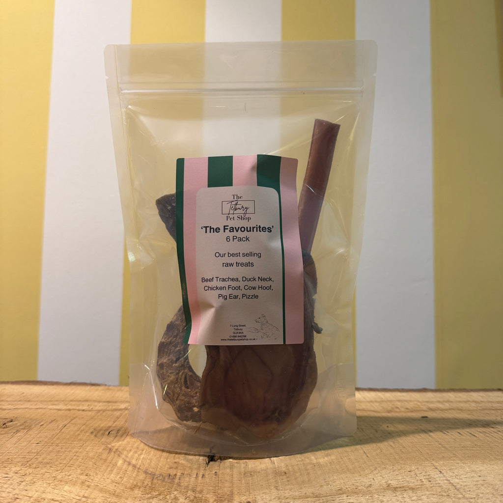 A packaged variety pack of raw dog treats, visible through the clear plastic, with a label showing different treats inside like beef trachea, duck neck, chicken foot, cow hoof, pig ear, and pizzle.